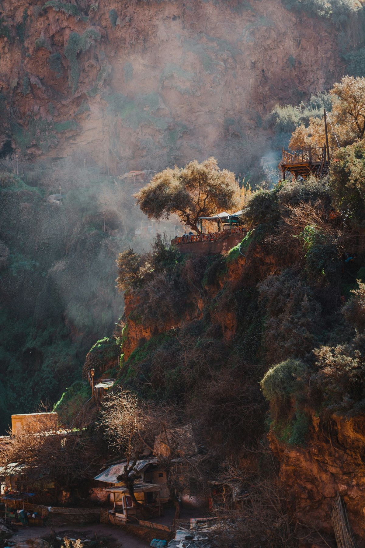 This image is a painting showing a cliffside village with houses built into a rocky, tree-covered hillside. The scene is bathed in soft, warm light, creating a picturesque and serene atmosphere.