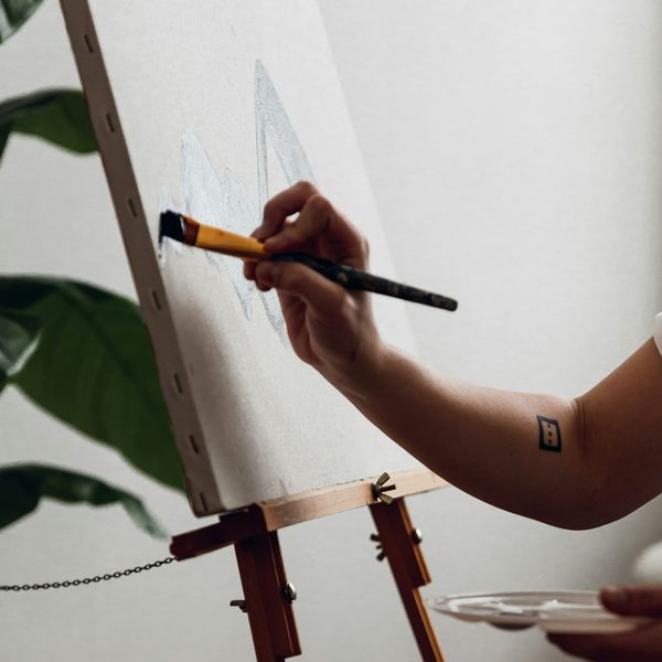 Artist painting on a canvas with a brush, holding a palette, in a bright room with a green plant and natural light, showcasing a creative workspace.