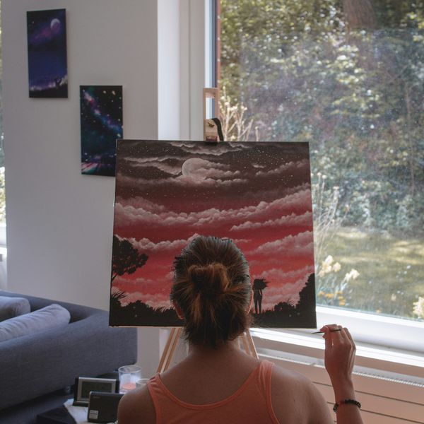 Artist painting a vibrant sunset landscape with a full moon and clouds on a canvas, seated by a window with a view of greenery, in a cozy living room setting.